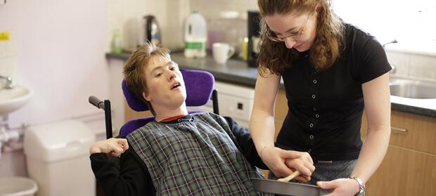 Two women in a kitchen. A young female carer supporting another woman in wheelchair with cooking equipment.