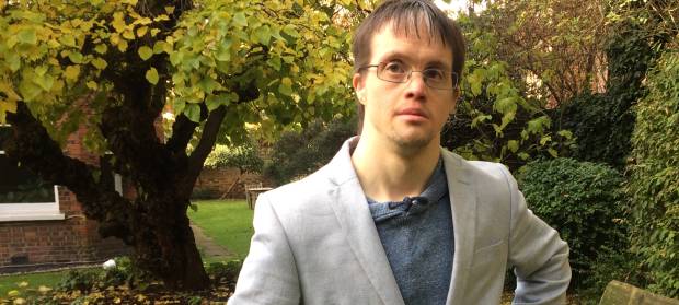 Man with glasses wearing pale jacket stood outside with trees and grass in background.