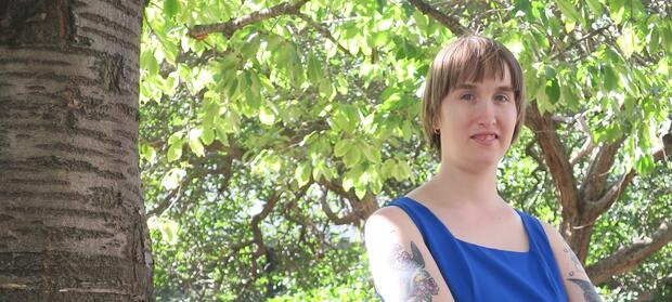 Young woman with short brown hair and blue top stood outside under a tree.