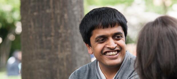 Young man smiling sat outside in front of tree.
