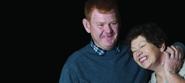 Man and woman smiling stood against black background. The man has his arm around the woman's shoulders.