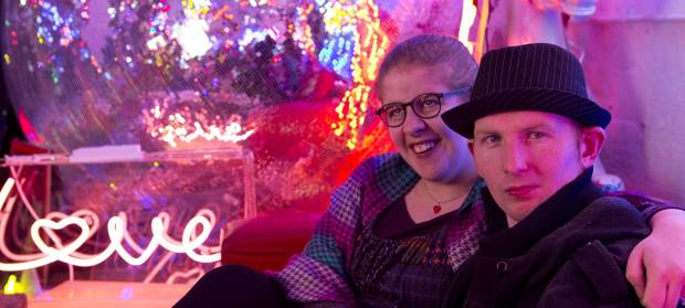 Man and woman sat next to each other on sofa it brightly coloured room, neon sign saying "love" in background