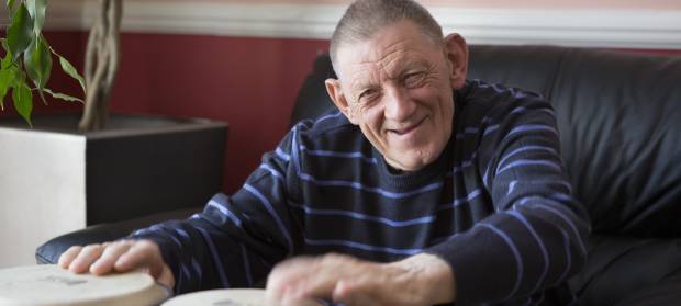 Smiling older man wearing navy blue sweater sat on black sofa playing with bongo drums