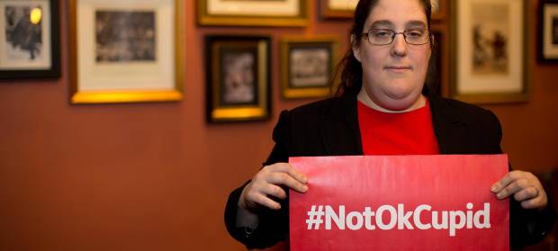 Woman stood in room with picture frames hung behind her, holding a sign that reads "#NotOkCupid"