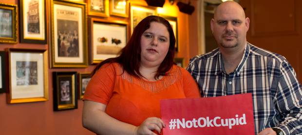 Man and woman stood in room holding a sign reading "#NotOkCupid"