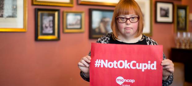Woman with red hair and glasses stood in pub room, holding a red sign with white writing that reads "#NotOkCupid"