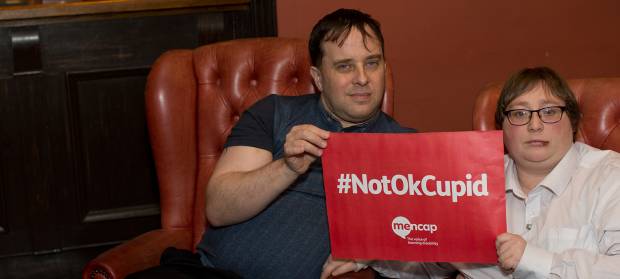 Man and woman sat on red sofa, holding a sign saying "#NotOkCupid"