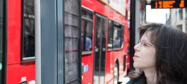 Woman with long dark hair and glasses stood at bus stop where red bus is stopped, looking at timetable.