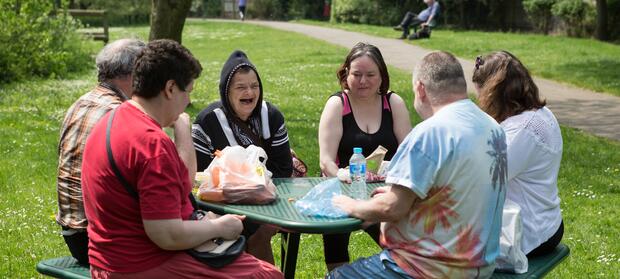 Group of people sat around picnic table in park, eating lunch.