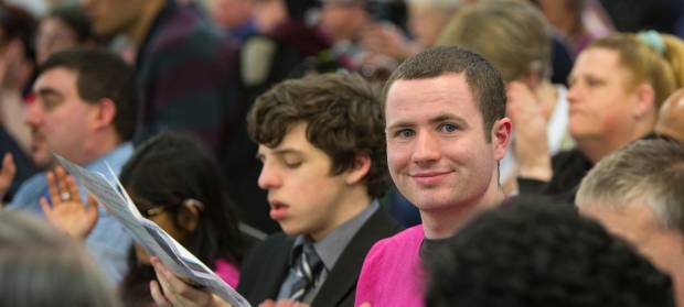 Young man with brown hair wearing pink Mencap t-shirt smiling and looking into camera whilst in crowd of people.