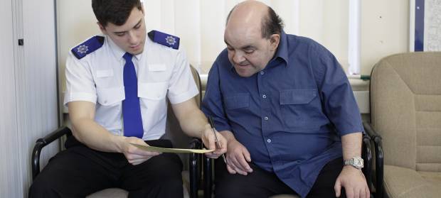 A policeman and man wearing navy shirt sat next to each other on chairs, looking over a piece of paper that the policeman is holding.