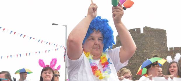 Woman wearing large bright blue wig, holding a colourful instrument, dancing in parade