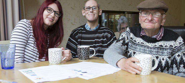 A woman and two men sat at table with cups of tea.