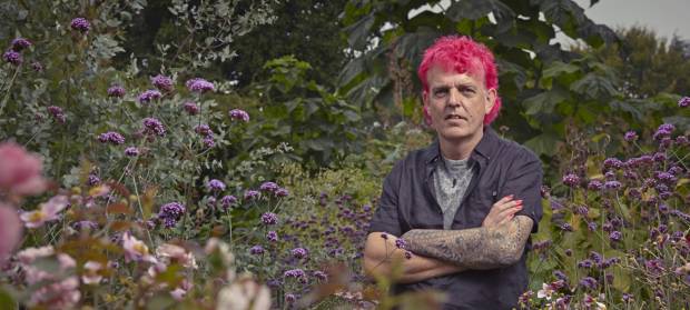 Man with pink hair stood in garden with arms folded