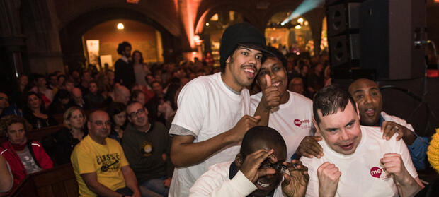 Group of 5 people huddles together smiling and posing whilst wearing white Mencap t-shirts, crowd of people in the background