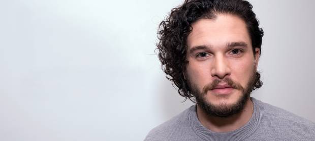Man, Kit Harington, with dark curly hair stood against white background looking into camera