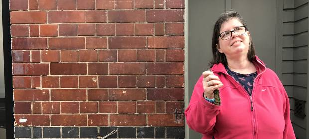 Smiling woman wearing pink fleece holding keys in her hand whilst stood in front of brick wall
