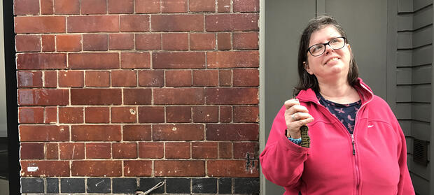 Smiling woman wearing pink fleece holding keys in her hand whilst stood in front of brick wall