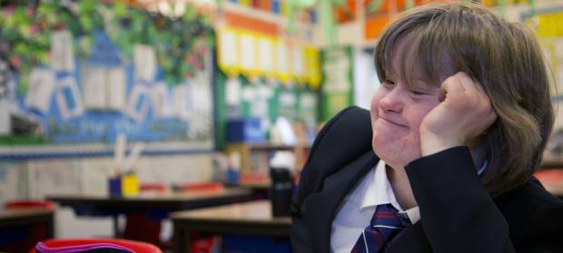 Schoolgirl sat in classroom smiling whilst resting her had on her hand.