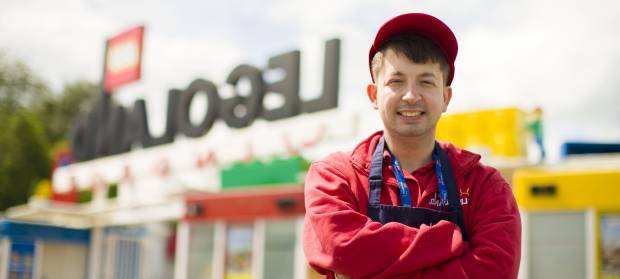 Young man wearing Legoland employee uniform stood in front of Legoland sign.
