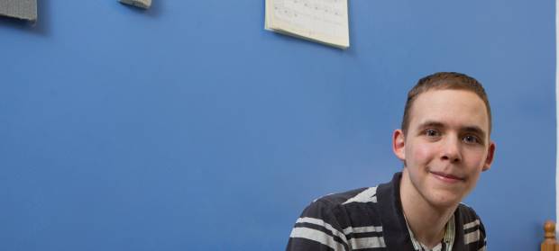 Young man smiling and looking into camera whilst sat in front of blue wall in bedroom.