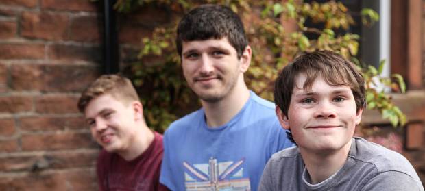 Three young man smiling into camera whilst sat outside house.