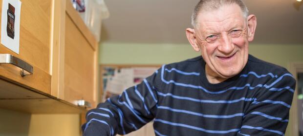Older man with short grey hair and blue striped jumper stood in kitchen smiling into camera.