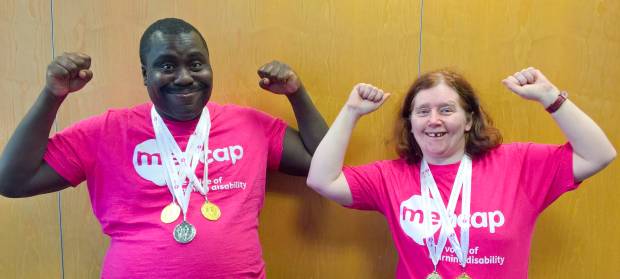 A man and woman stood next to each other with their arms raised. Both are wearing Mencap t-shirts and have medals round their necks