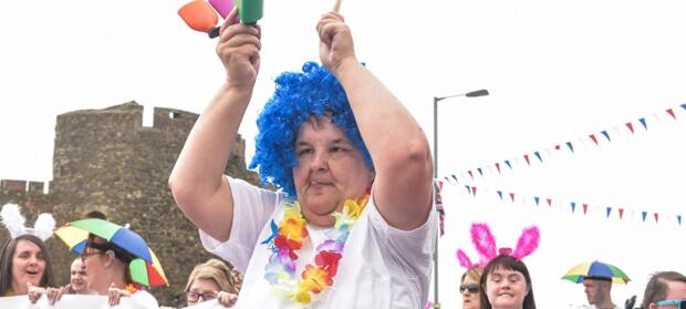 Woman wearing large bright blue wig holding colourful instrument taking part in a parade