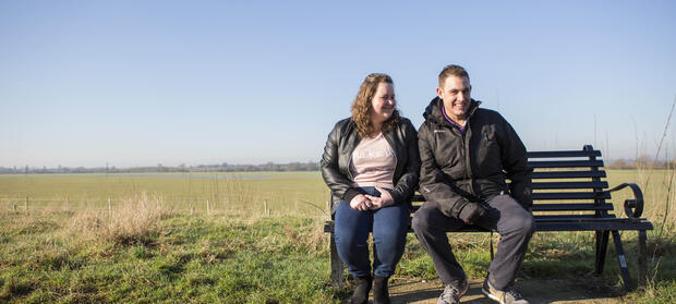 Man and woman sat smiling on bench in field.