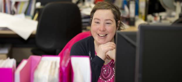 Woman with short brown hair sat at her desk in an office smiling.