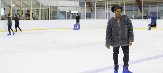 Young woman smiling at camera wearing fluffy coat, jeans and ice skates stood on ice in ice rink.