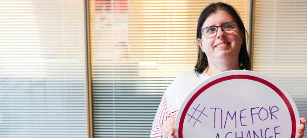 Woman with dark hair and glasses stood in office holding a sign that reads #TimeForAChange