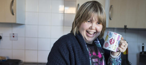Woman with light hair stood  in kitchen. She is holding a mug near her face and is smiling.