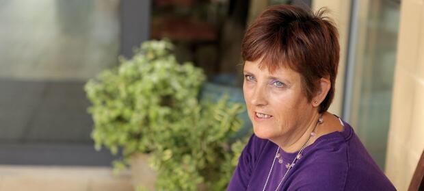 Woman with short hair wearing a purple top is sat outside next to plant