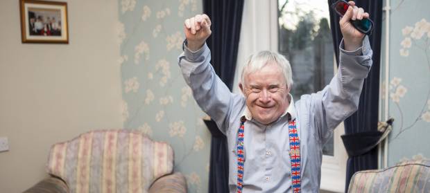 Smiling older man with shirt and Union Jack braces stood in living room raising his arms in the air in celebration.