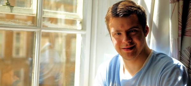 Young man wearing blue top smiling into camera whilst sat next to window.