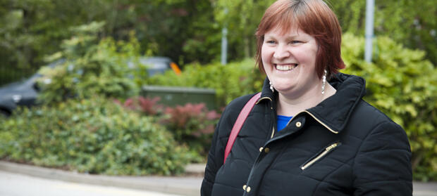  Woman stood outside smiling and wearing a black overcoat. Bushes and a car in the background.