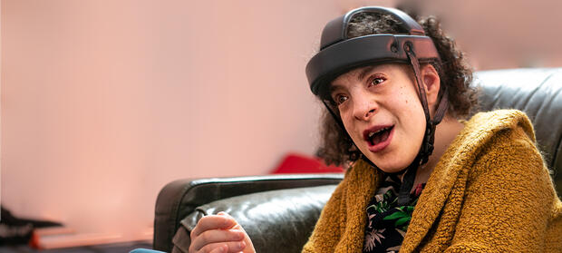 Young woman with dark curly hair wearing a yellow jumper sat smiling on black sofa in front of pink wall.