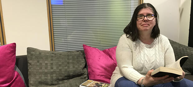 Woman with dark hair and glasses wearing a white knitted jumper is sat on a grey sofa with pink cushions. She is smiling and looking into the camera whilst holding a book in her hands.