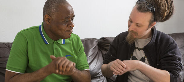 Two men sat together on black sofa doing exercises with their hands.