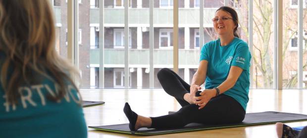 Woman sat on a toga mat on floor smiling whilst doing a yoga pose