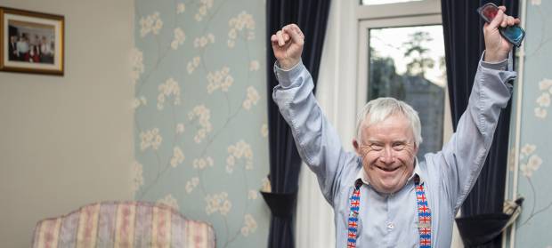 Older man stood in living room smiling with his arms raised in the air in celebration
