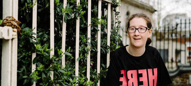 Young woman with brown hair and glasses sat outside in front of white railings, smiling into camera.