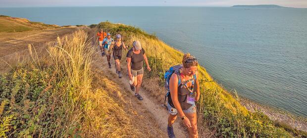 Five people walking in single file along a coastal path with sea in the background.