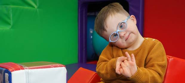 Young boy wearing blue-rimmed glasses and mustard coloured jumper. He is sat in colourful soft play area holding his hands together. His head is titled to the side and he is looking into the camera.