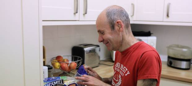 A picture of a man wearing a red t-shirt. He is stood in a kitchen, he is smiling as he opens a packet of bread.