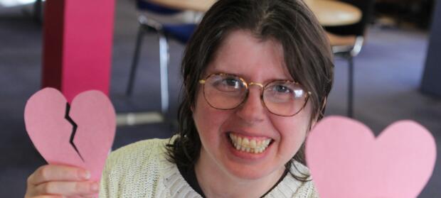 Woman with short dark hair and glasses smiling whilst holding pink paper hearts.