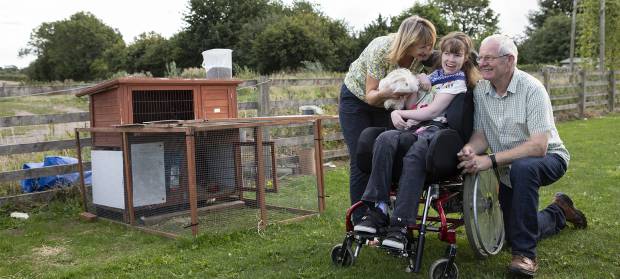 Mother and father stood with their daughter who is using a wheelchair. They are in a garden next to an animal pen, the mother is holding a guinea pig for her daughter to look at.