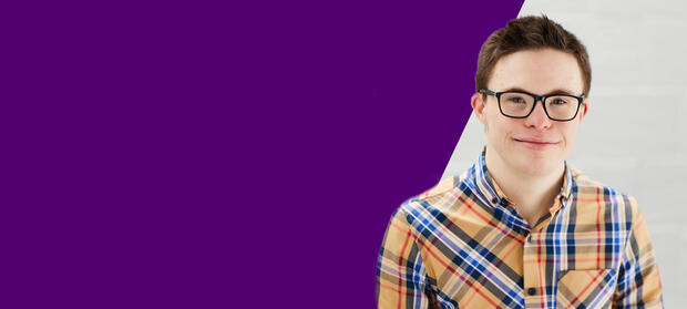 George has short dark hair, a yellow plaid shrt and glasses. He looks at the camera in front of a white background and smiles.